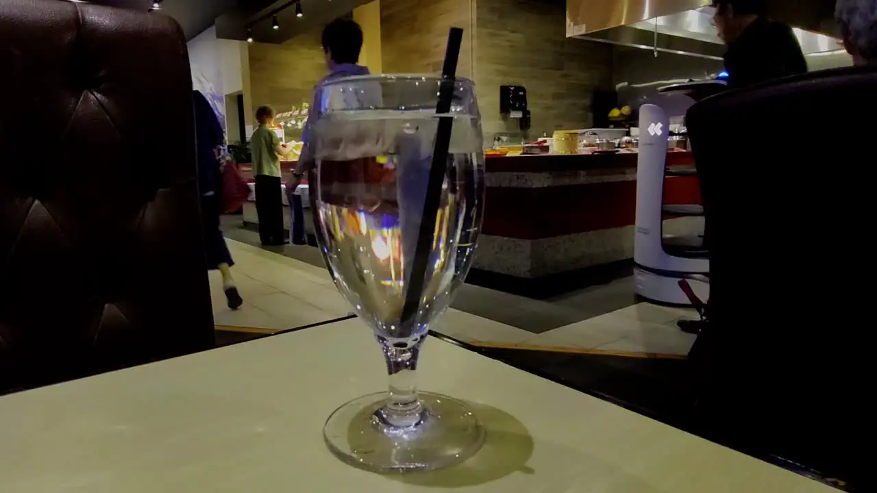 closeup of clear glass cup with water and black straw in the foreground while background AI robot directs people to seating area at fast food eatery buffet all you can eat multicultural people place