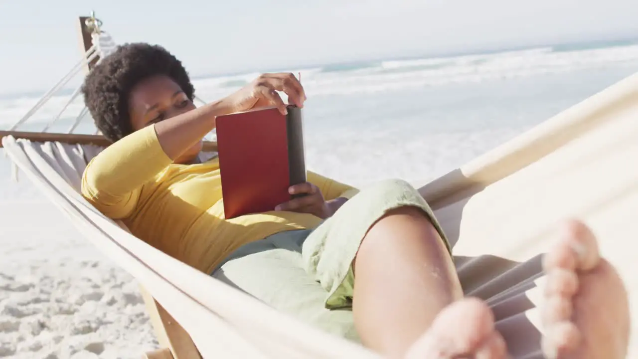 Happy african american woman reading and lying in hammock on sunny beach