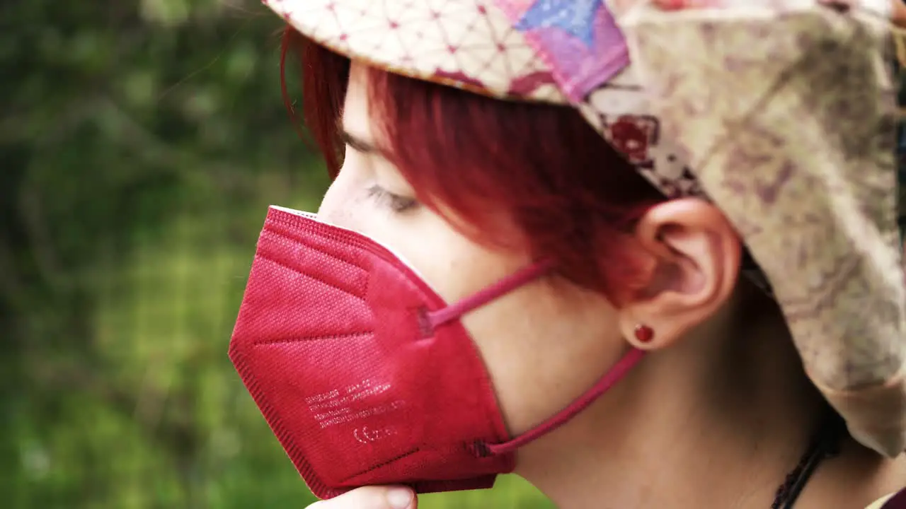 Side view of a red haired girl covering her face with a medical ffp2 mask in a green outdoor environment