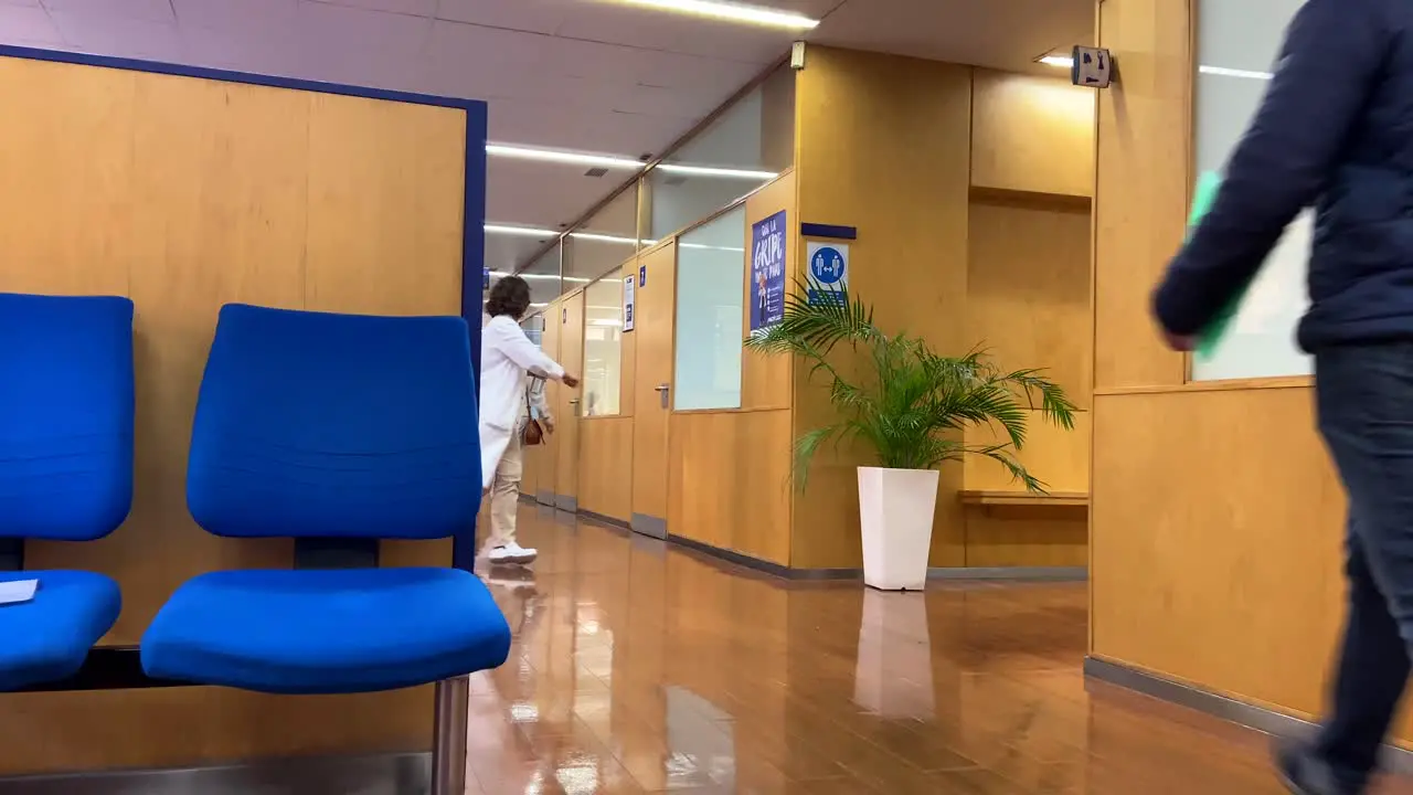 Older doctor walking in white coat and mask down the hallway of a medical clinic from the waiting room