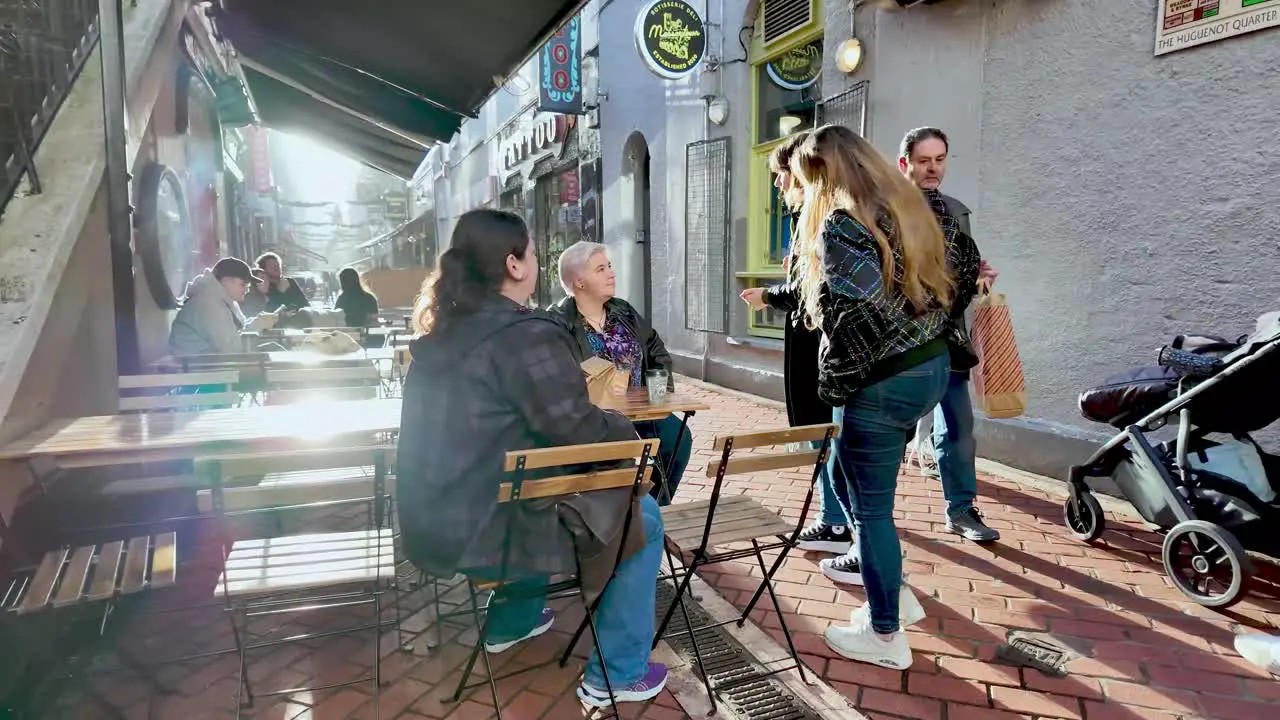 Early morning sun on the street in Cork City and group of young women sitting on a pubs terrace with pedestrians passing in slow motion