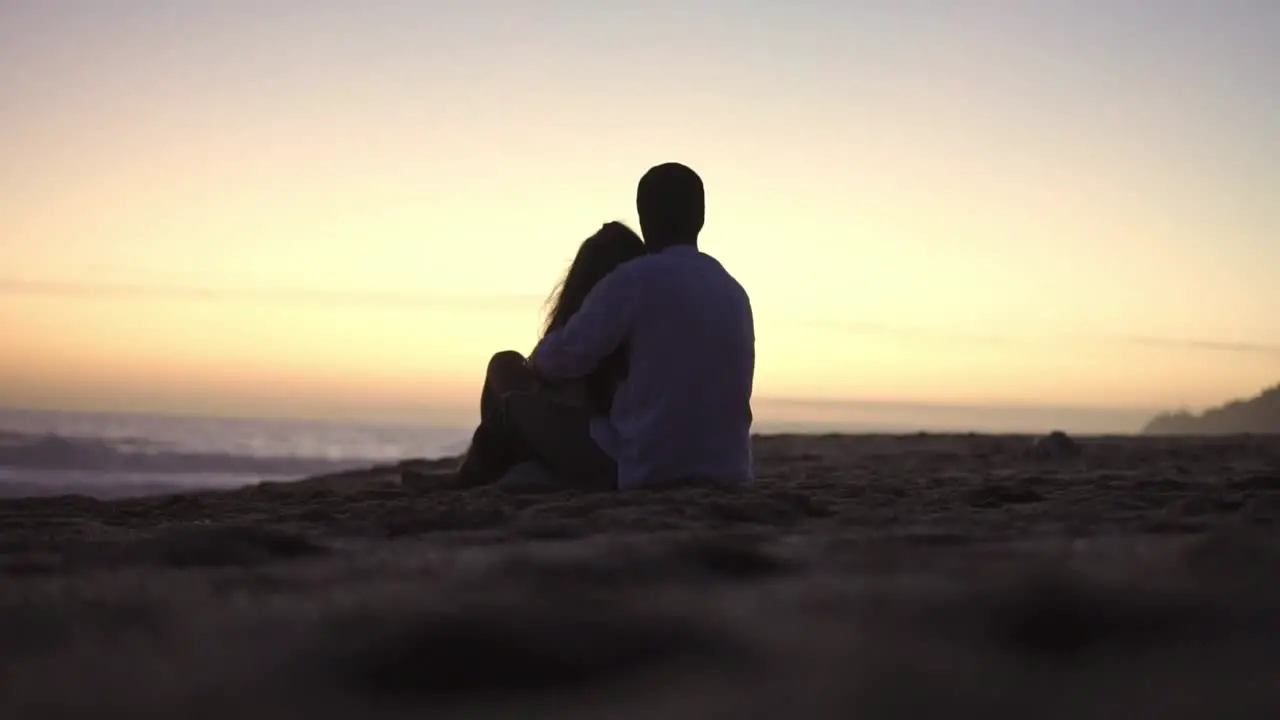 Static handheld shot of a couple in love in silhouette sitting on the beach in front of the sea with calm waves during a romantic sunset