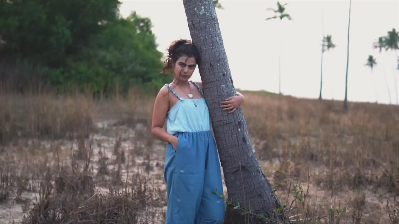 A young girl in a blue dress is holding a tree on an overgrown beach and looking at the camera