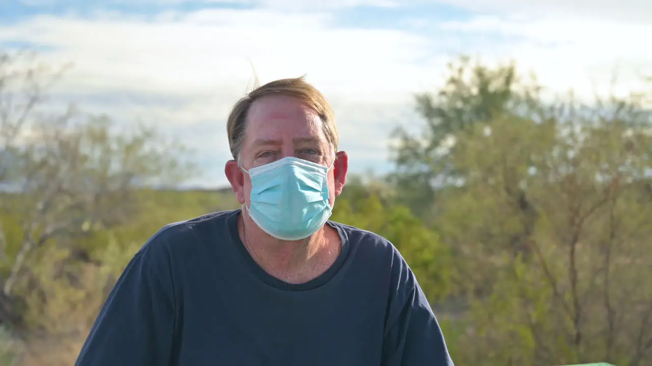 Senior adult man outdoors in public park wearing medical face mask looks at camera