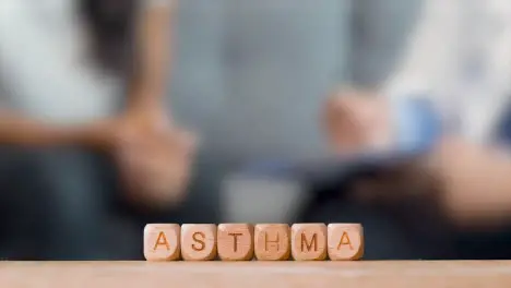 Medical Concept With Wooden Letter Cubes Or Dice Spelling Asthma Against Background Of Doctor Talking To Patient