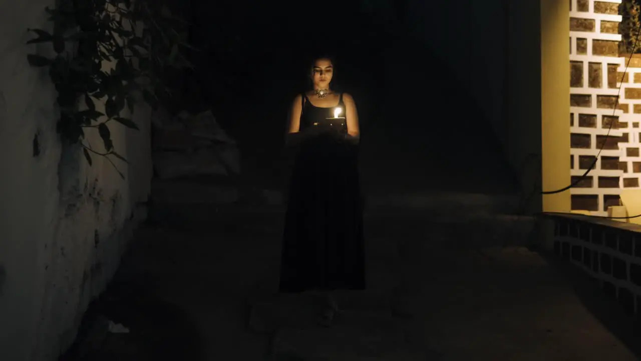 Front view of woman in black dress walks down the dark stairs holding a burning candle in her hands with bushes nearby