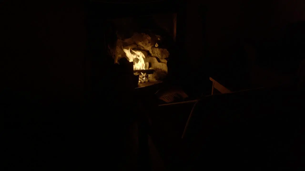 An old chair is illuminated in a dark room by the light from a log fire in an old fireplace