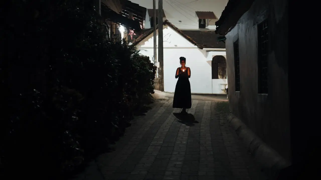 Front view of pretty woman in black dress walking along dark street over mosaic floor holding lit candle in hands while dog looking at her in background