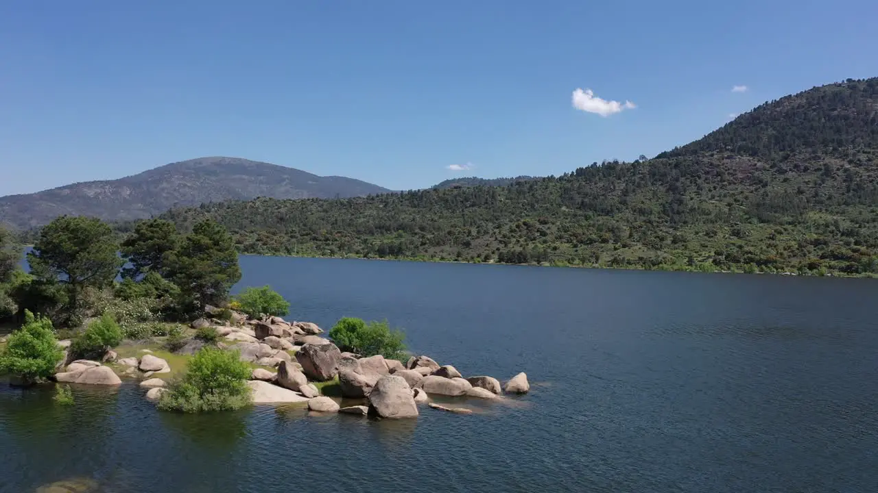 flight with a drone in a reservoir full of water with mountains and forests seeing on the left a small island with vegetation and granite stones on a sunny afternoon and a blue sky in Avila-Spain