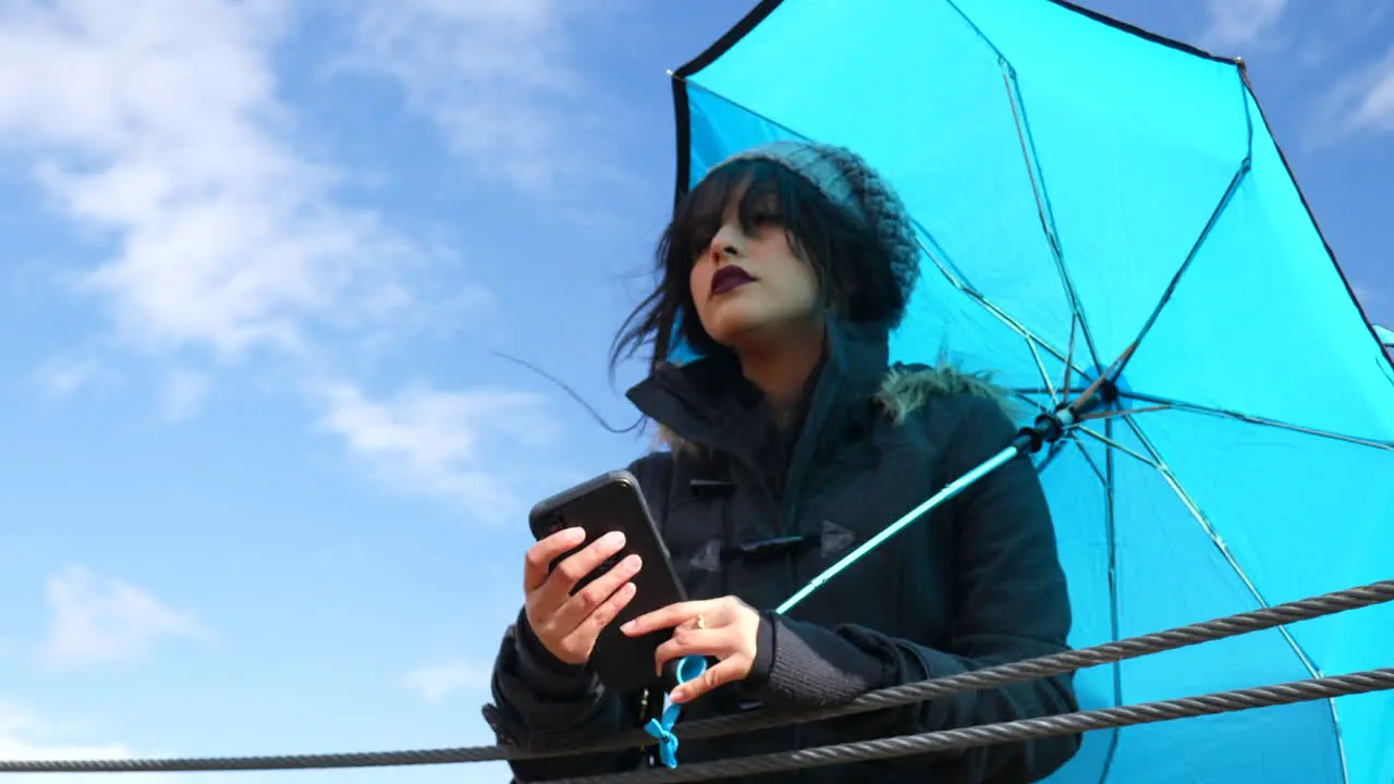 Pretty young woman checking the weather or texting on smartphone with umbrella under blue skies as rain storm clouds approach