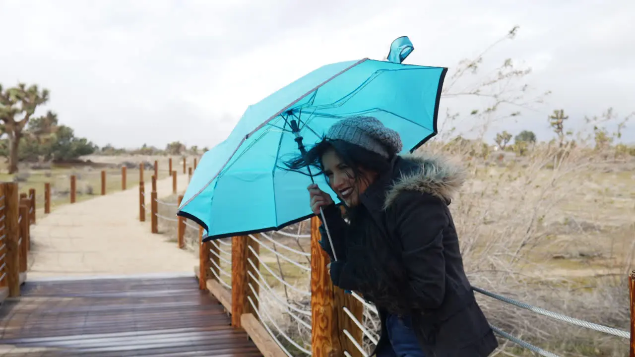 A beautiful woman in bad weather with a blue umbrella during the strong winds of a rain storm in the desert SLOW MOTION