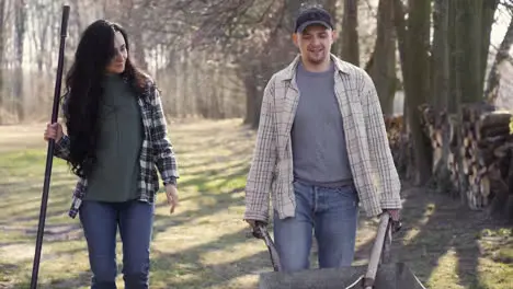 Caucasian couple and their dog holding a wheelbarrow and a rake while they are walking throught the countryside