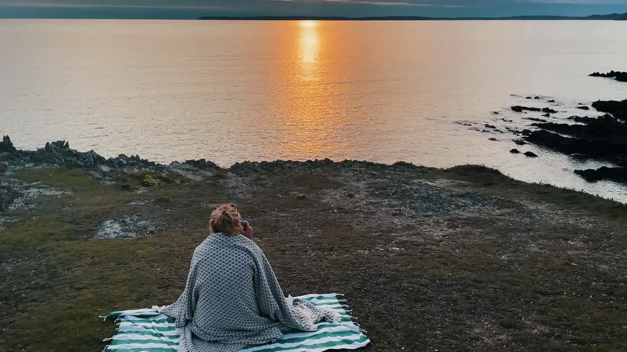 Aerial of young hipster woman sitting on blanket in front of Atlantic Ocean in France Brittany taking photo during sunset golden hour