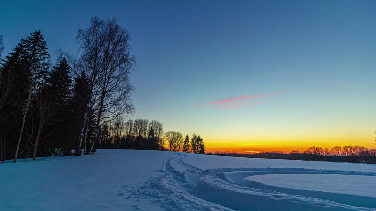 Cinematic sunset timelapse at horizon through snow covered landscape with silhouette fir trees and tyre print in foreground