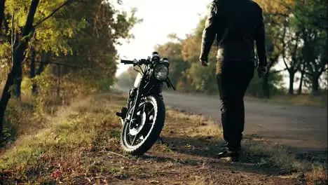 Unrecognizable man in helmet and leather jacket coming up to his bike and starting the engine while standing on the roadside in a sunny day in autumn