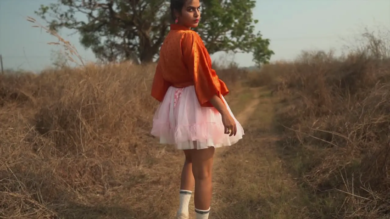 Girl in a red shirt and a pink skirt walks towards a tree and looks back into the camera as she's walking