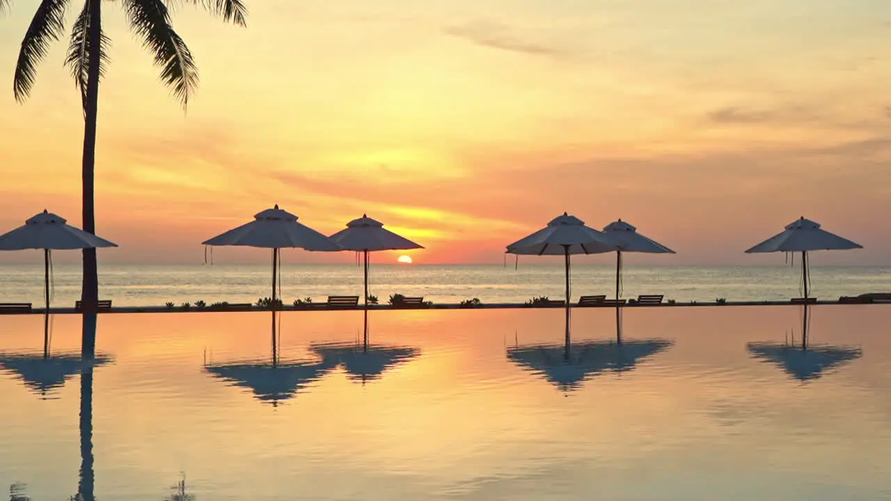 Orange Tropical Sunset Above Sea and Pool of Luxury Resor With Tree and Parasols Silhouettes and Mirror Reflection on Water Static