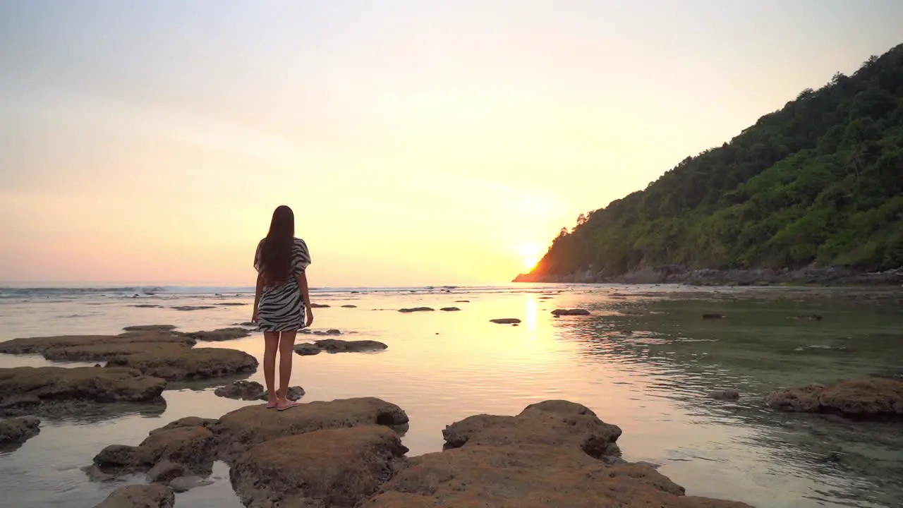 A young woman with her back to the camera stands on the exposed rocks from the low tide as she admires the colorful sun setting behind a mountain