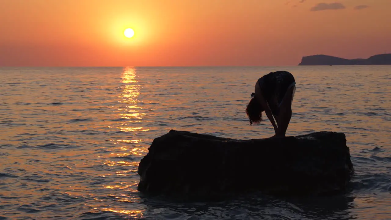 Young woman poses Yoga meditation exercises over big cliff washed by sea at beautiful sunset