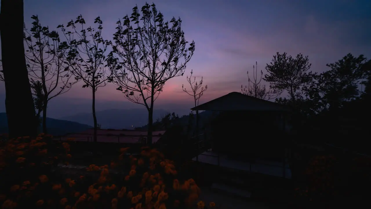 Evening timelapse of sun setting down followed by lights glowing up in a valley with a foreground of Flowers trees and a tent-house