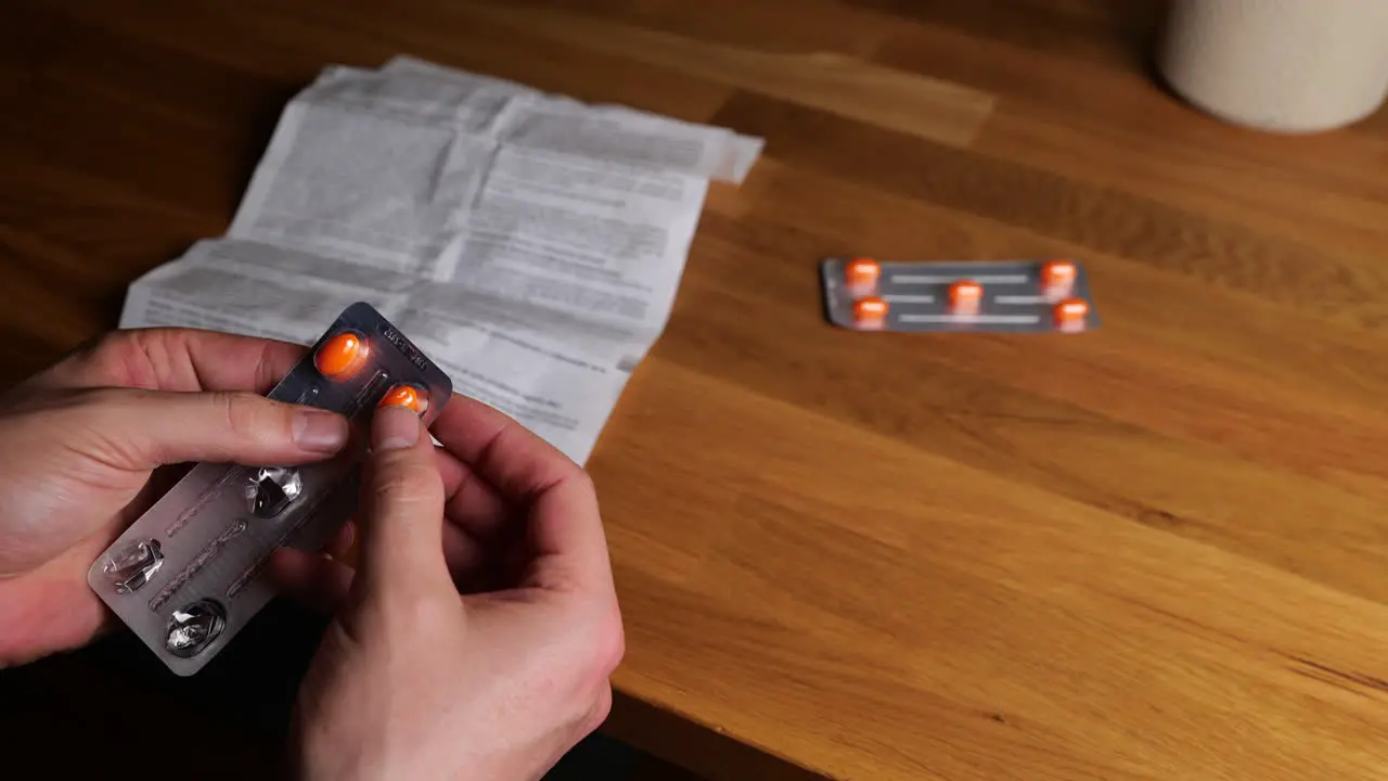 Hands holding blister pack of orange medication tablets with person reading medicine guide sheet at wooden table