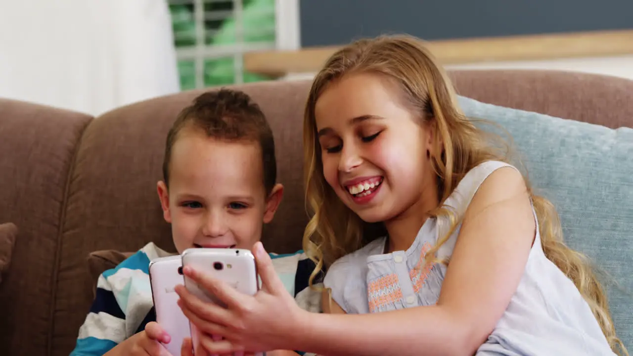 Smiling boy and girl sitting on sofa and using mobile phone