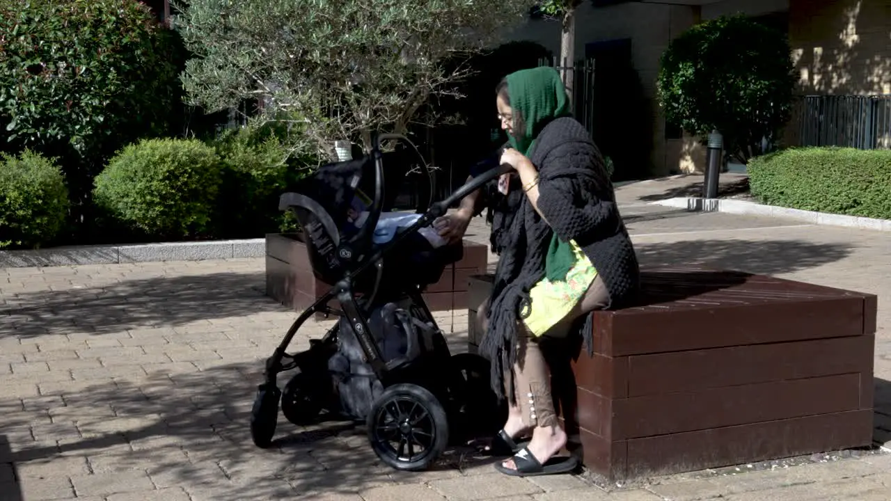 A young Asian Muslim mother comforting her newborn baby in a pram while sitting in a park outdoors on a sunny day