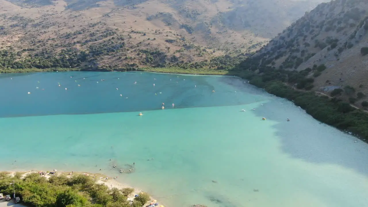 Drone view in Greece flying over a light and dark blue lake with small boats and surrounded by green mountain on a sunny day in Crete