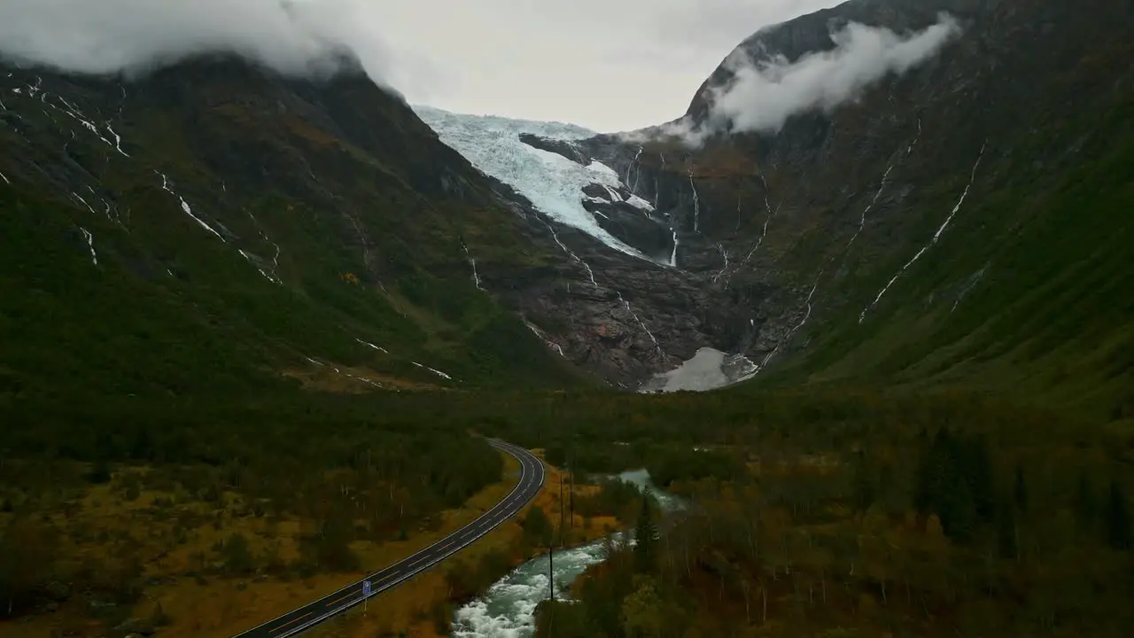 Drone flies through a valley over a Norwegian road towards a cloud-covered Gletcher