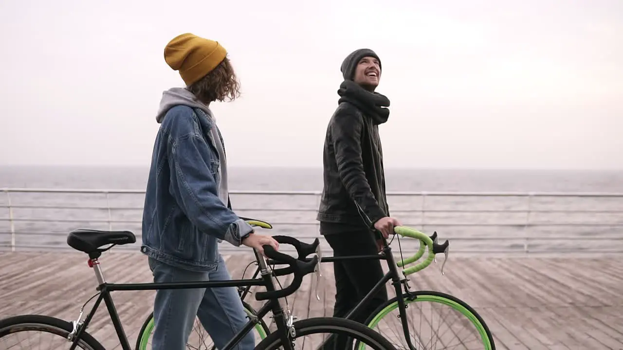 Side View Of Smiling Couple Of Young Hipsters Walking Together With Their Bikes Near The Sea At Autumn Day