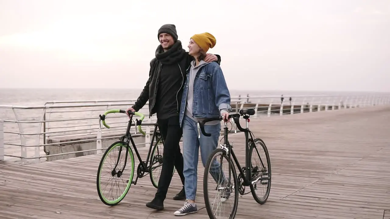 Full Length Of Couple Of Young Hipsters Walking Together With Their Bikes Near The Sea At Autumn Day