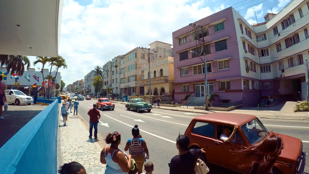 Footage of Havana Cuba on a sunny and windy day with people waiting and walking on the sidewalk with usual traffic driving up and down the avenue