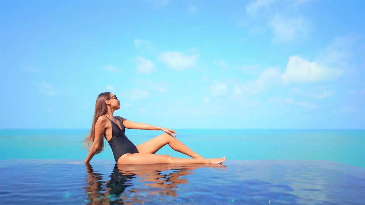 Sexy exotic female in swimsuit sitting on infinity pool edge enoying in stunning view of tropical sea horizon slow motion