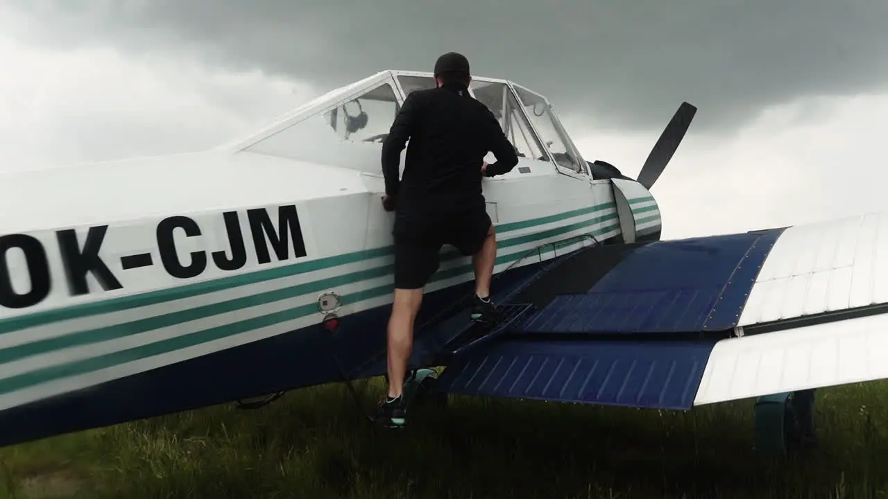 Following FHD shot of a man climbing on a wing of a small sport airplane preparing to enter the cabin and take off from a field airport with a grey stormy sky above