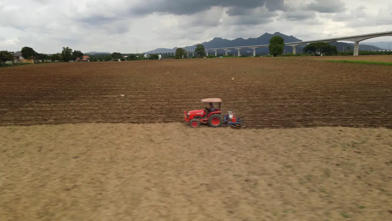 Sliding towards the left aerial footage of a hard working farmer driving red tractor across farmland tilling and preparing for new batch of plants high-speed railway network on horizon