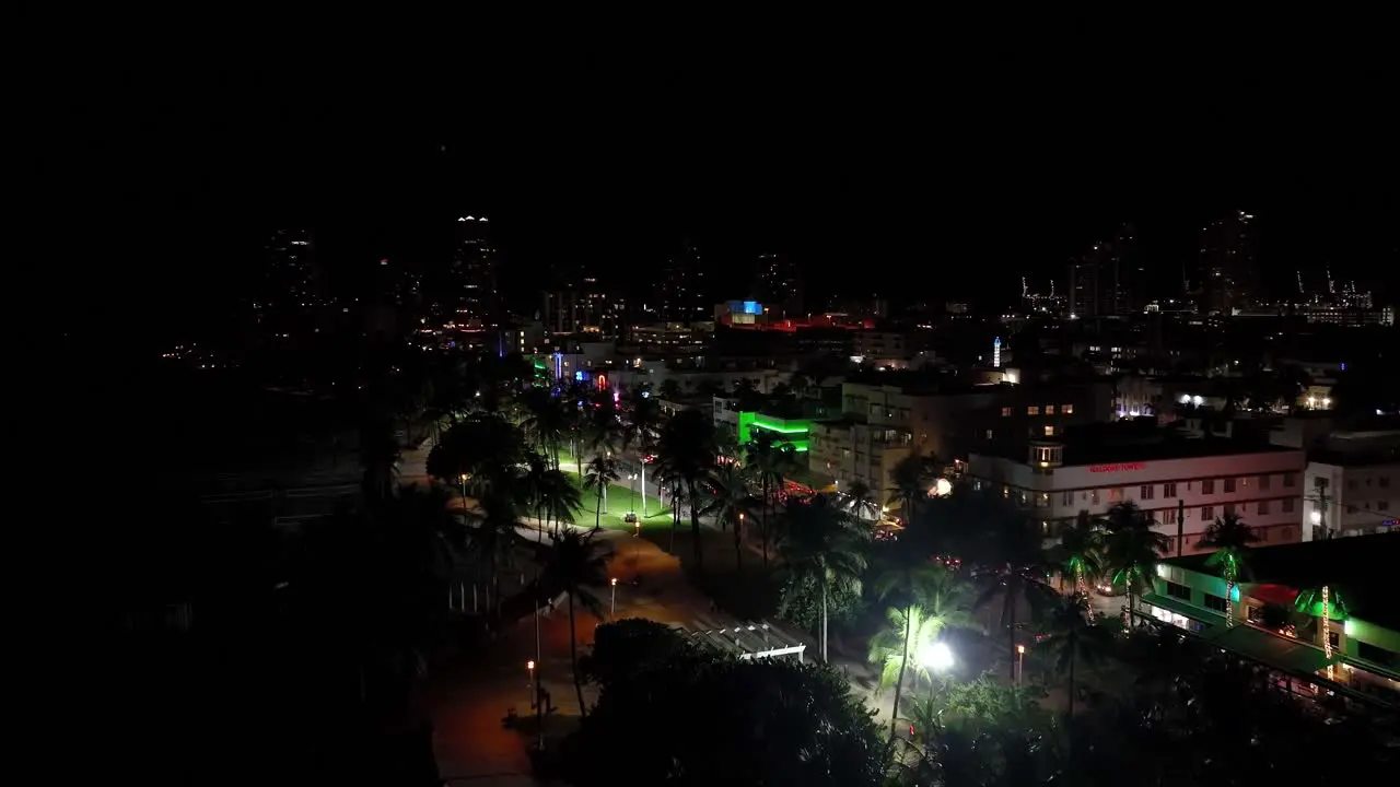 Nightly aerial drone footage of Miami USA forward movement showing many different colored lights over the buildings together with palm trees