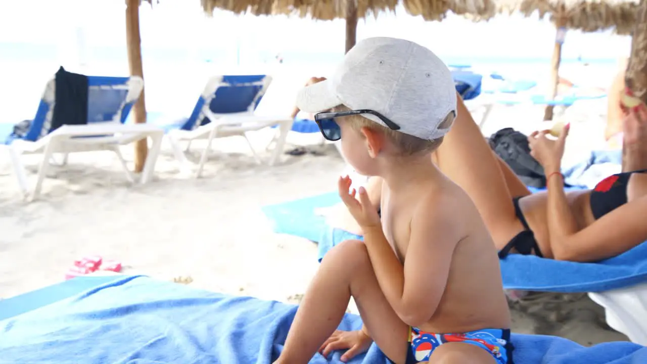 Child sitting on the sunbed at the beach plays with his hat and sunglasses removing them and putting them several times