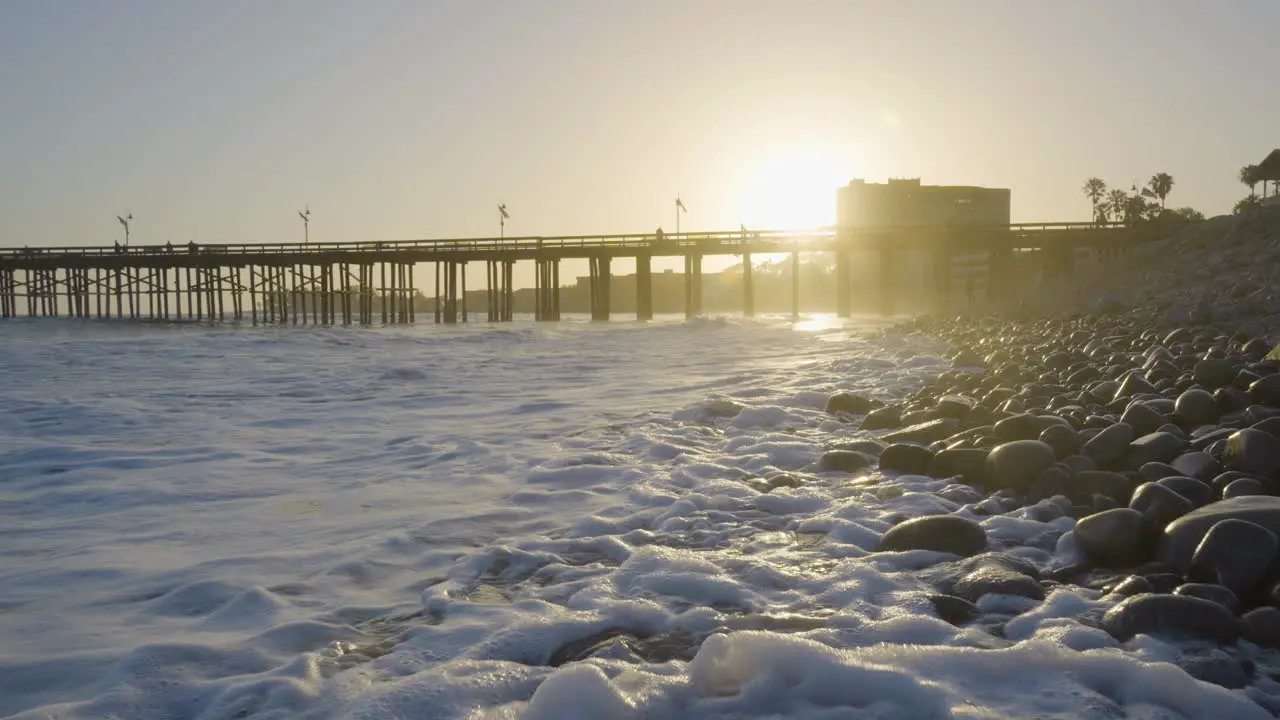 Dolly shot moving back of the Ventura Beach with waves crashing along the rocky shores with the Ventura Pier in the background at sunset located in Southern California