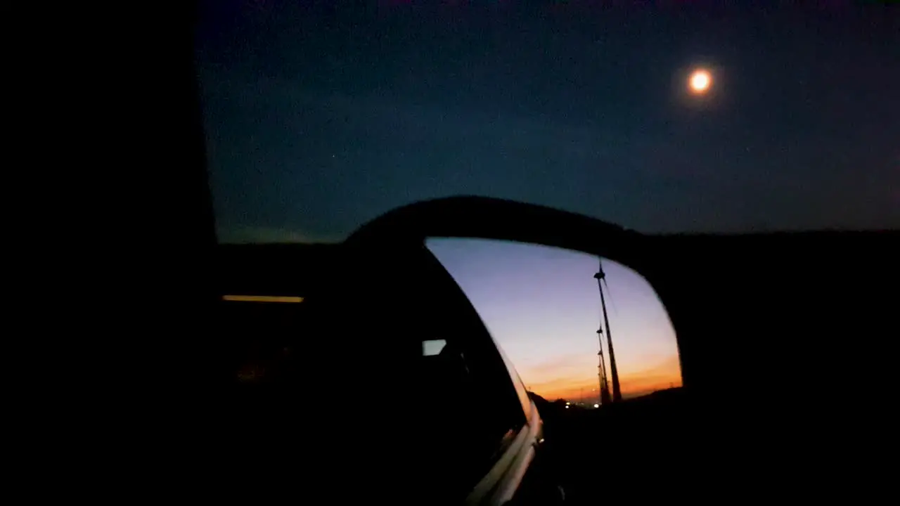 Long row of wind turbines seen through rear-view mirror with hazy moon in the background during dusk while driving