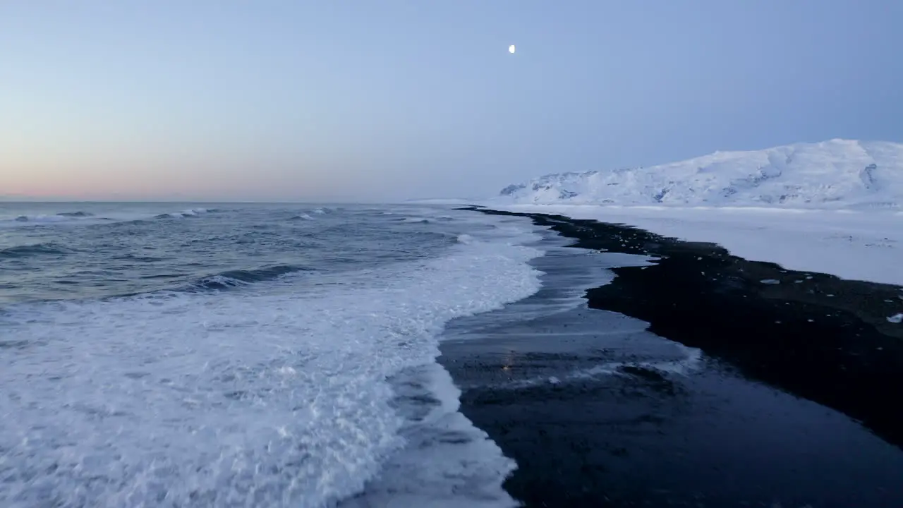 AERIAL Flying over Black Beach with white arctic snow mountains in background in Iceland in Winter Snow Ice Waves Water