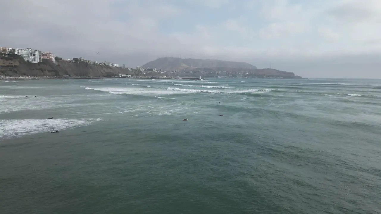 Slow aerial view dolly shot in front of the coast in miraflores in peru of the turquoise sea with calm waves and surfers in the water with view of rocks buildings and mountains in the background