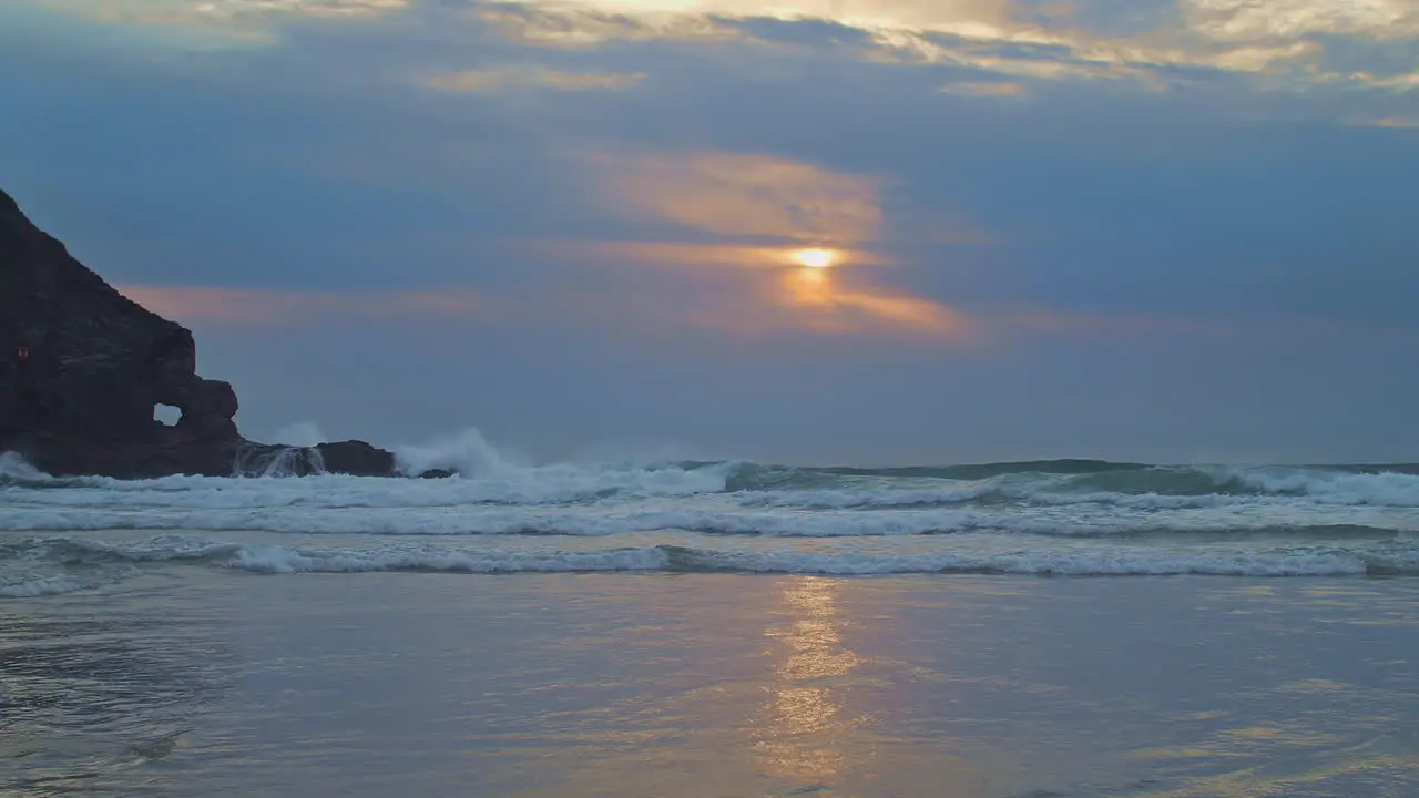 Surfers ride evening waves as glowing sun fades into clouds over beach