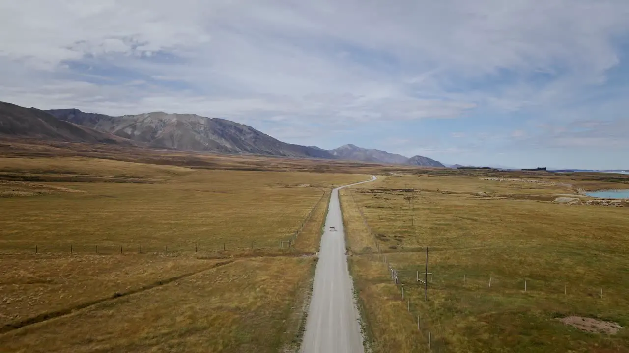 A vehicle navigating a rough dirt path surrounded by barren rural landscapes with majestic mountains and a tranquil lake in the distance bathed in warm sunlight