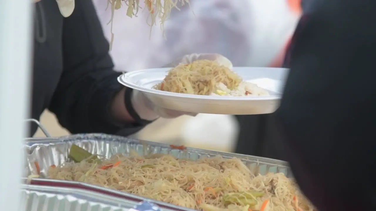 Person serving fresh hot noodles on white plastic plate with tongs outdoor buffet