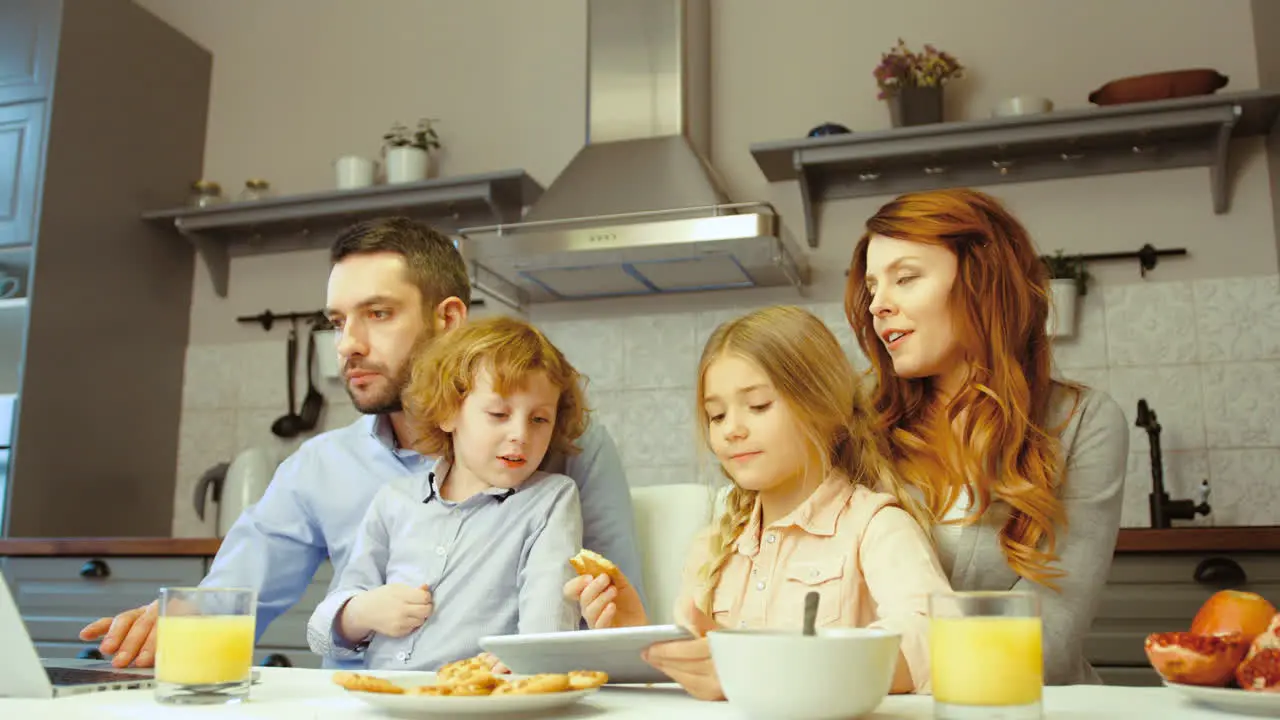 Cheerful Family Having Breakfast While They Use A Laptop And A Tablet