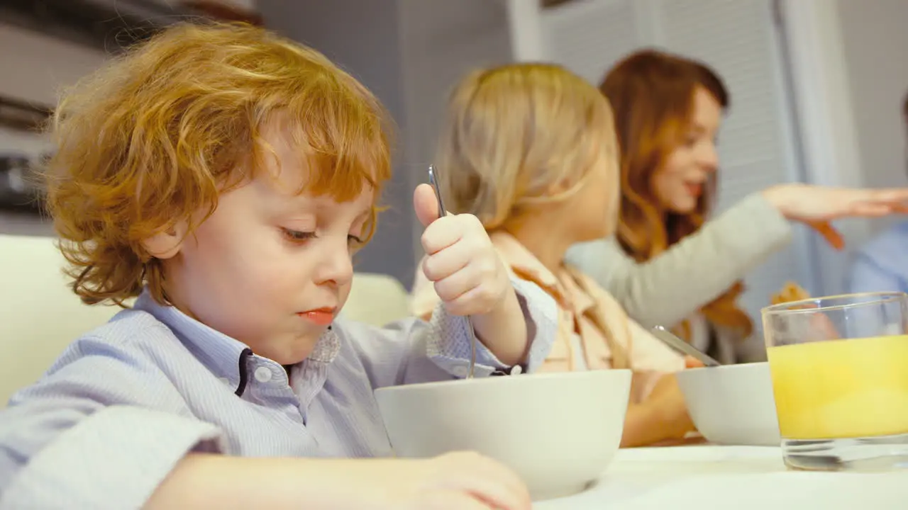 Lovely Little Boy With Blue Eyes Having Cereal While The Rest Of The Family Talks In The Kitchen