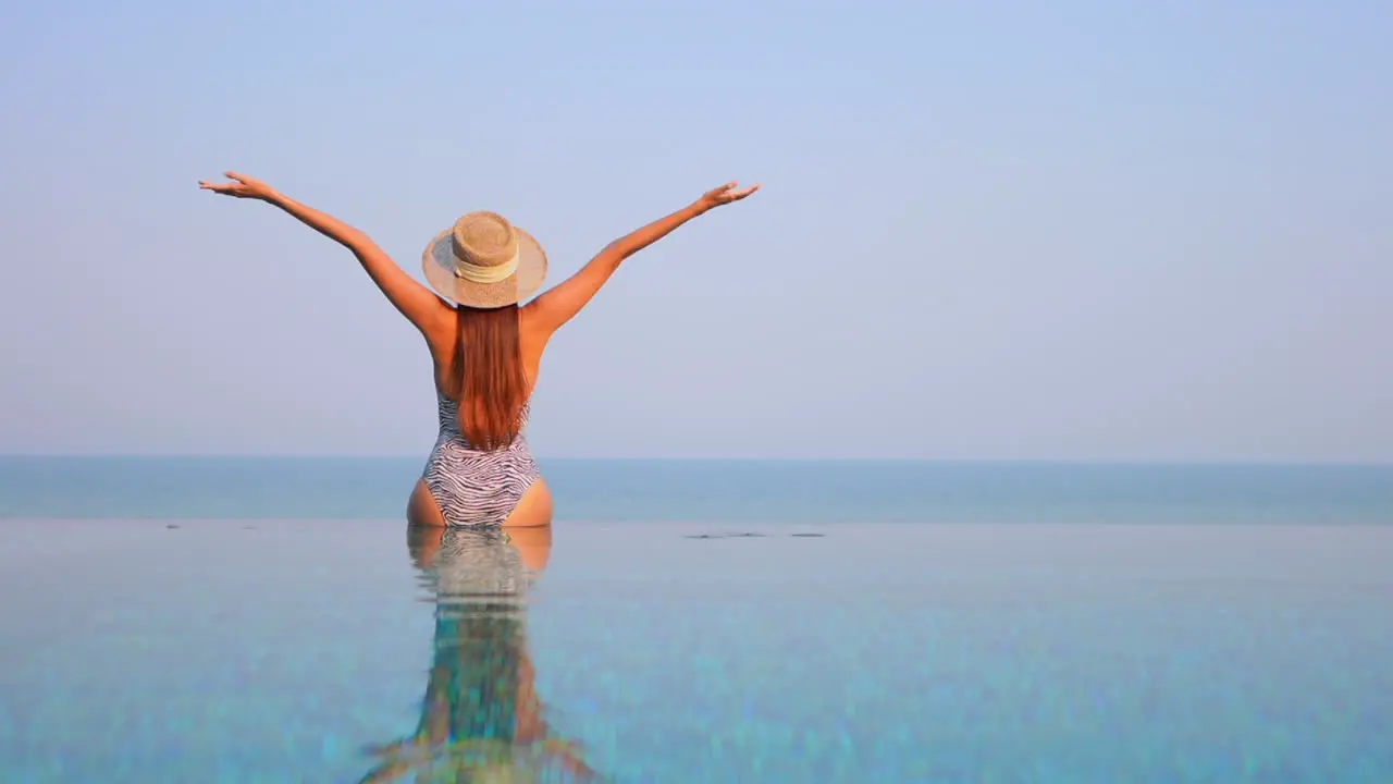 Back of exotic woman sitting on infinity pool border and raising hands tropical happiness on vacation slow motion full frame