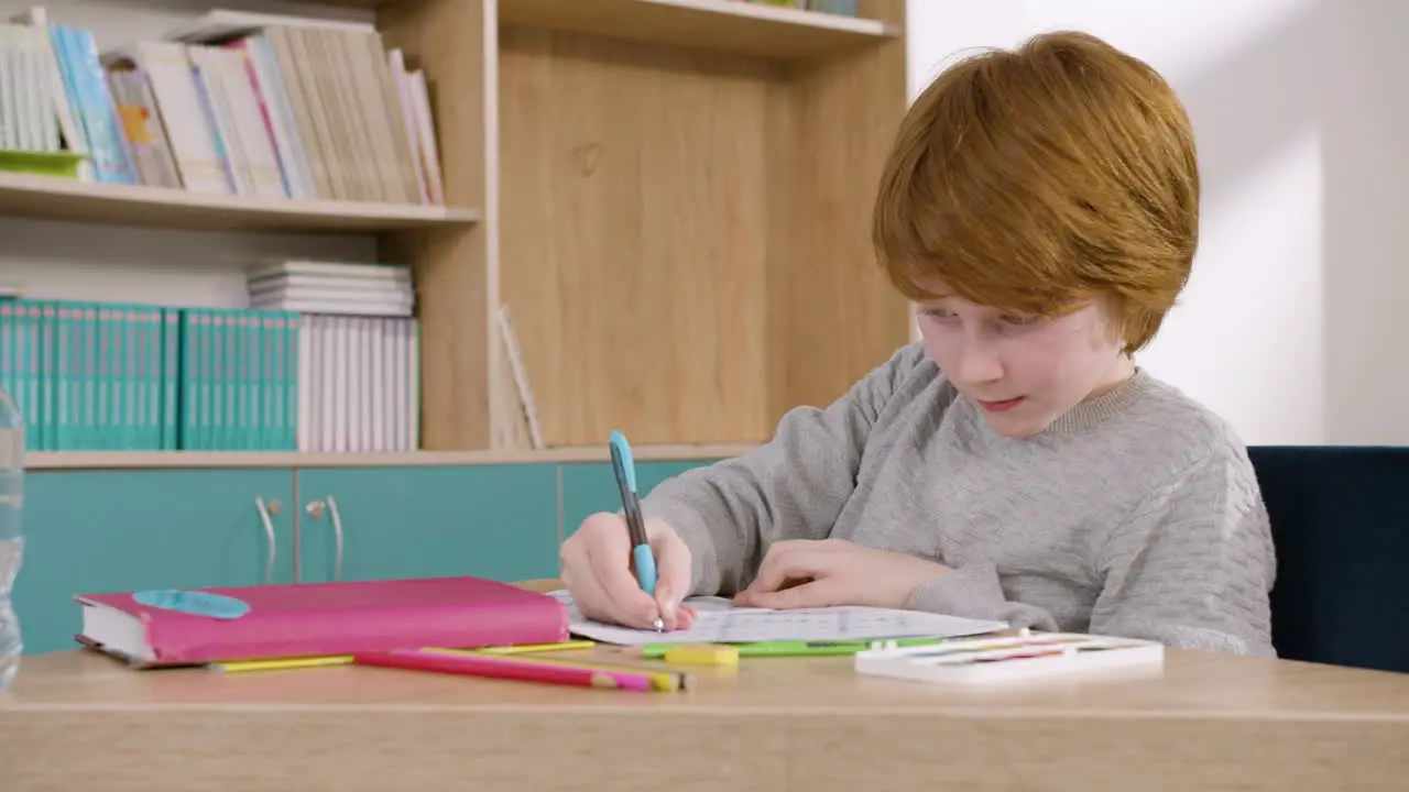 Ginger Boy Sitting At Desk And Drawing In Notebook During Class At School