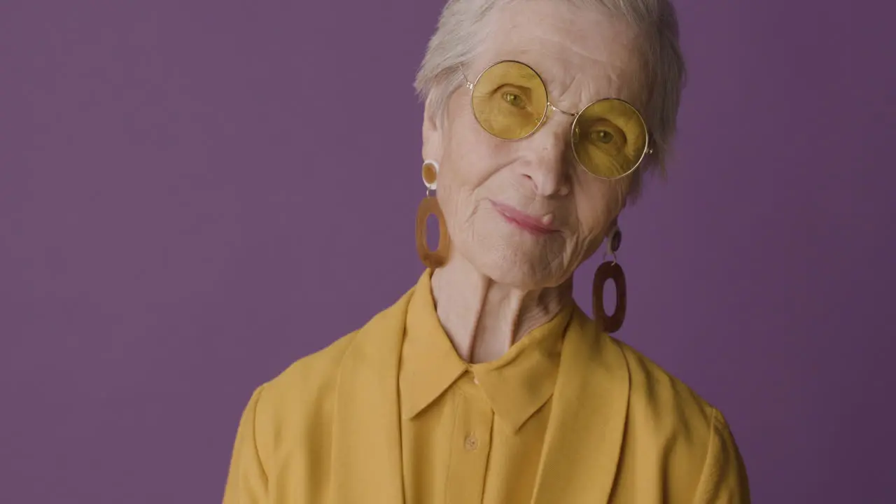 Senior Woman With Short Hair Wearing Mustard Colored Shirt And Jacket Earrings And Sunglasses Posing And Looking At Camera On Purple Background