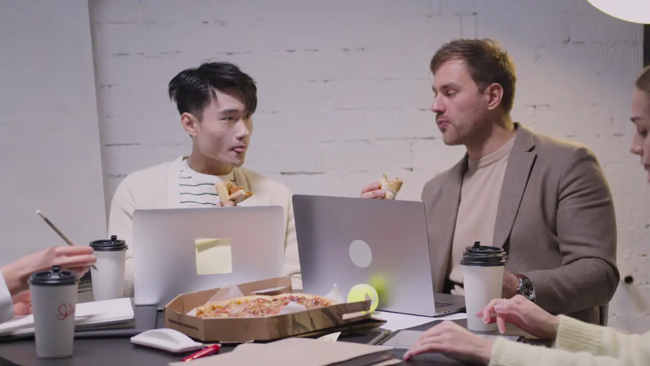 Two Multiethnic Colleagues Eating Pizza During A Team Meeting In The Boardroom